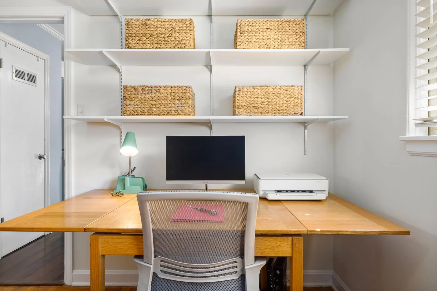 A clean, modern home office with a computer, shelves, and wicker baskets.