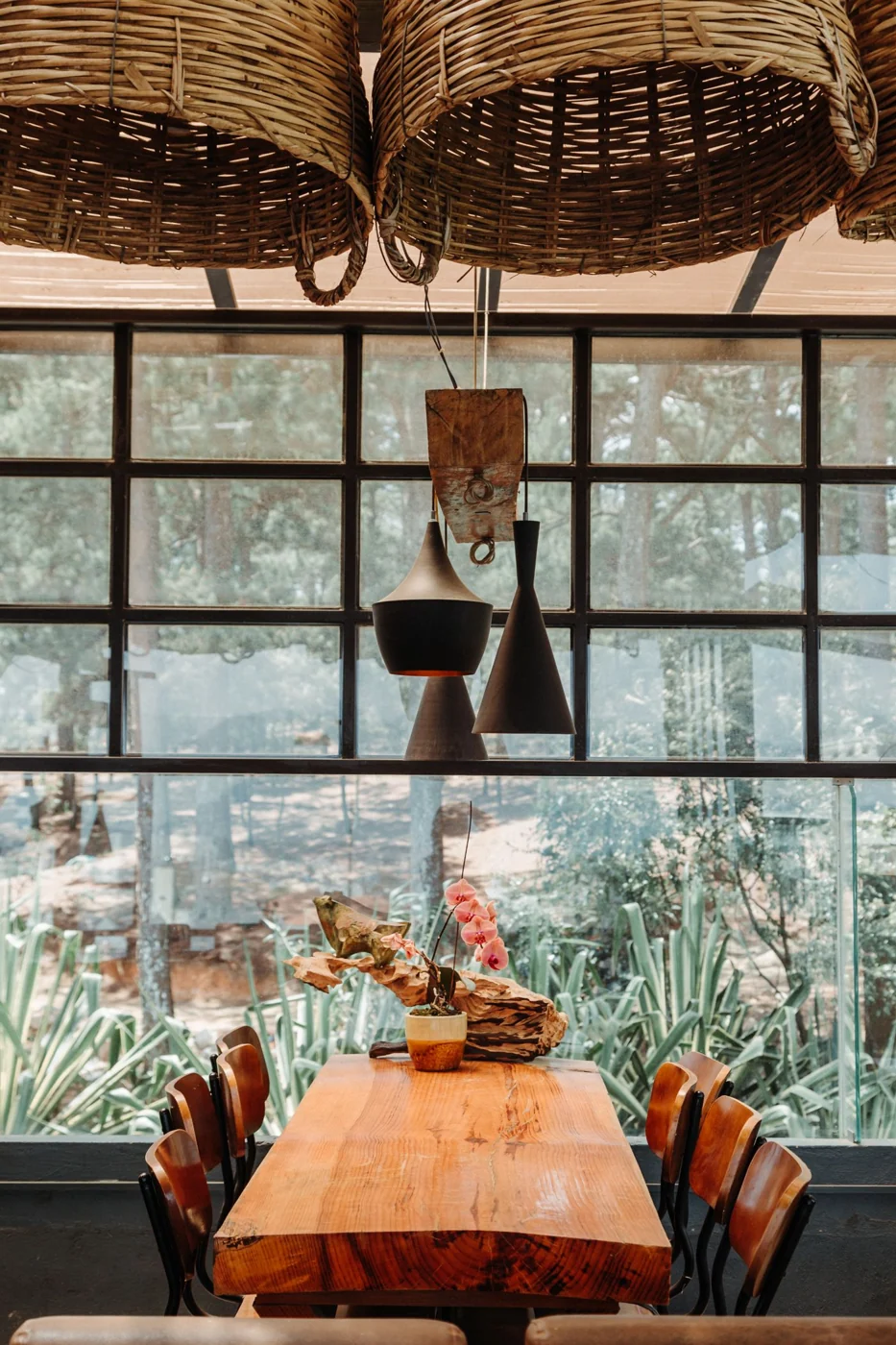 Cozy dining room featuring a wooden table, basket chandeliers, and a glass wall with a garden view.