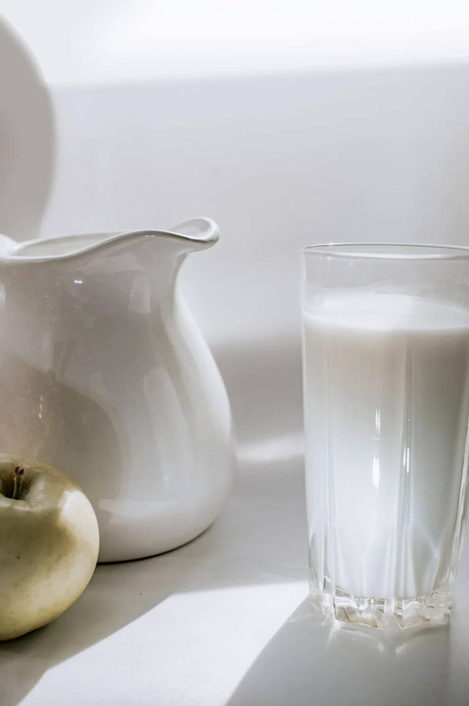 A serene still life image of milk in a glass next to a green apple and ceramic pitcher.
