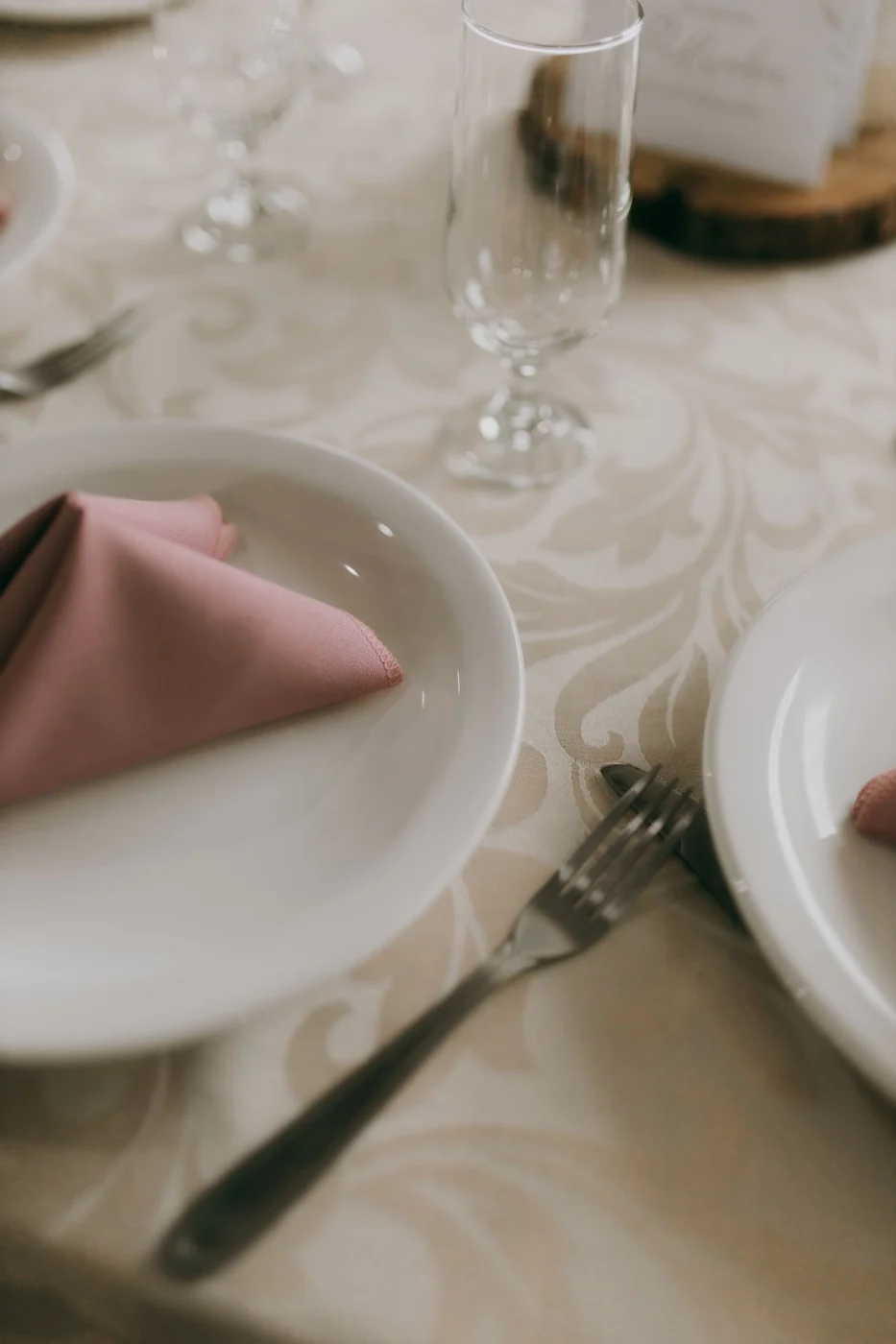 An elegant table setting with white plates, pink napkin, and glassware on a patterned tablecloth.