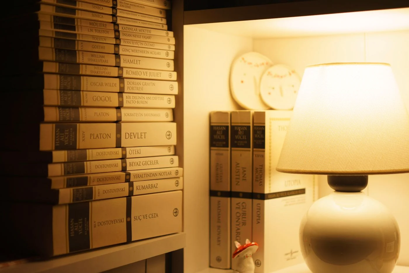 A warmly lit bookshelf with classic literature, a lamp, and decorative plates creating a cozy ambiance.