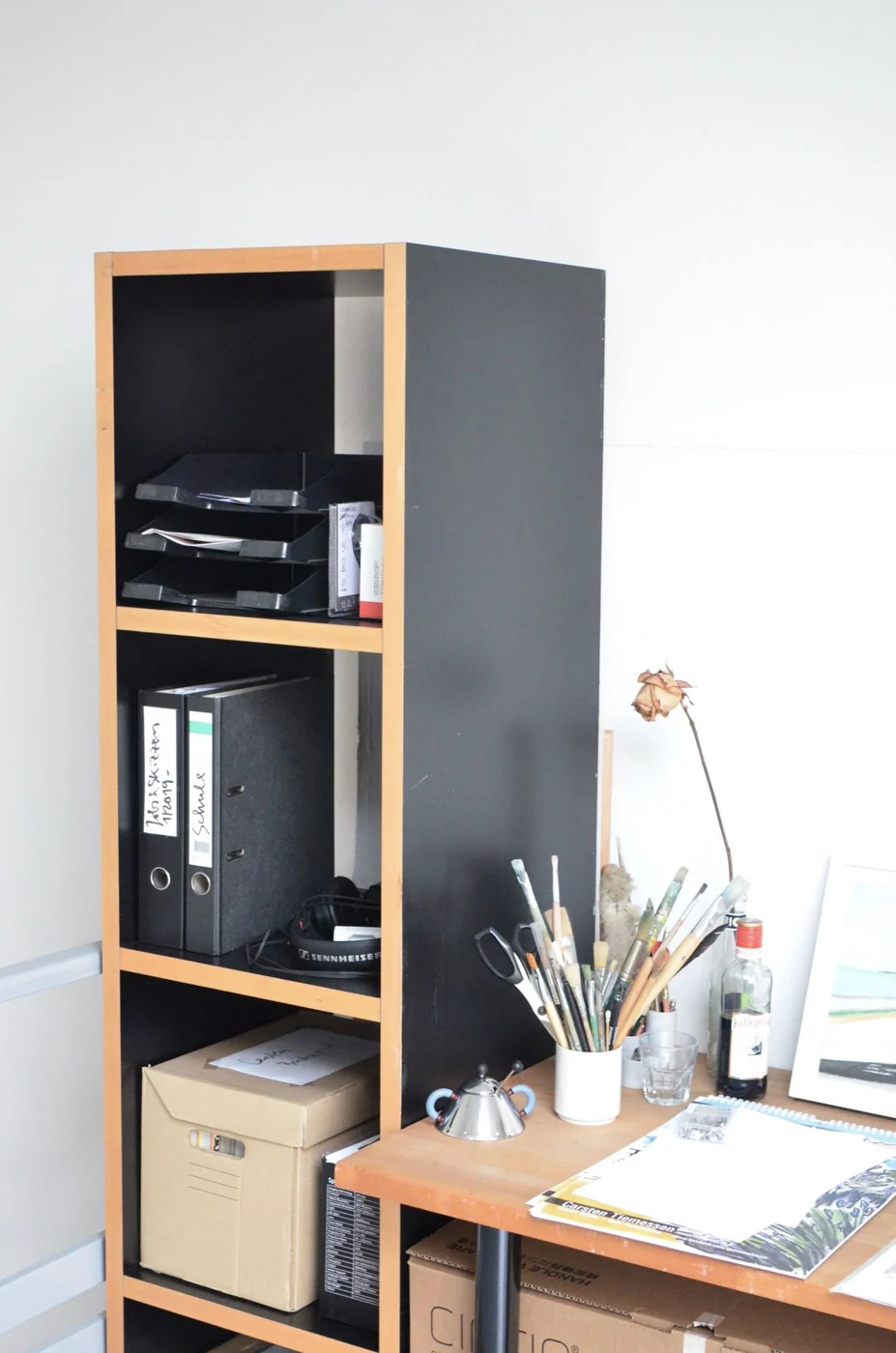A modern office workspace featuring a black shelf with documents and art supplies on a wooden desk.